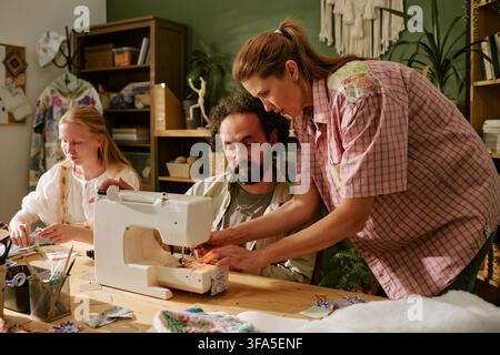 Woman Teaching Middle Aged Man Using Sewing Machine in Workshop Stock Photo