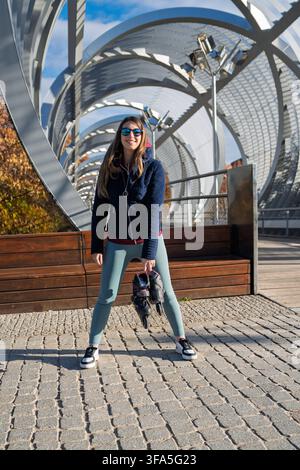 Smiling woman holding inline skates while standing on the modern urban bridge Stock Photo