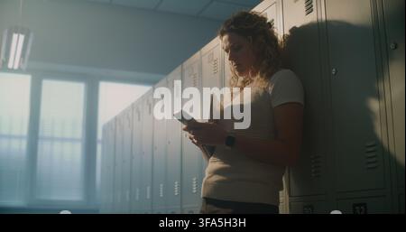 Female Teacher Standing Alone in Empty School Locker Room, Checking Smartphone Notifications and Responding to Messages. Concept of Modern Education, Digital Communication, Everyday Teacher Workflow. Stock Photo