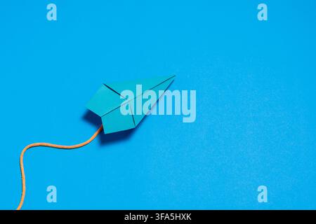 Blue paper airplane attached to an orange string placed on a bright blue background, representing imagination, creativity, freedom, travel Stock Photo