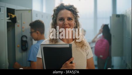 Portrait of Happy Smiling Female Teacher Holding Notebooks and Folders Standing in School Corridor with Lockers. Mentorship, Academic Support, Positive Communication in Modern School Environment. Stock Photo