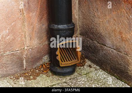 Close up view of a weathered black metal downspout with a rusty drain grate positioned against a textured brown stone wall on a tiled patio, industrial urban exterior architecture Stock Photo