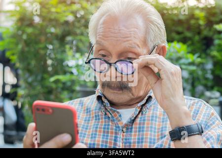 Asian elderly man sitting at park has problems with his eyesight for reading on his smart phone. Vision problems concept. Stock Photo