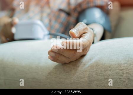 Elderly man checking his blood pressure at home using a digital monitor while sitting on sofa at home. Close-up, selective focus on hand. Stock Photo
