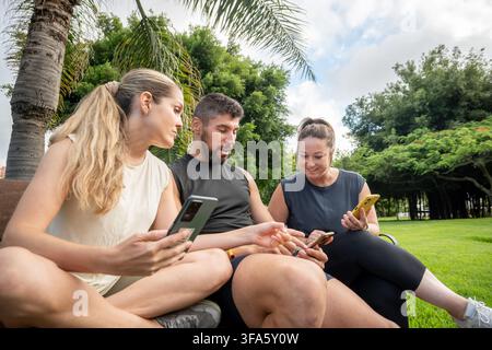 Group of friends sitting on a park bench using smartphones and talking together while taking a break after outdoor exercise. Stock Photo