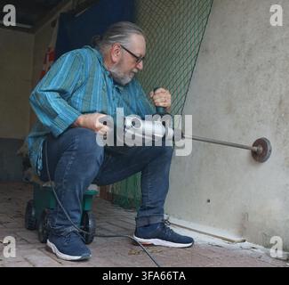 An older man, wearing glasses and a blue striped shirt, is sitting on a wheeled cart while using a jackhammer to bore into a brick wall. He is positio Stock Photo