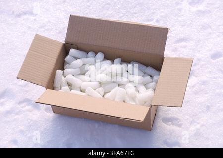 An open brown cardboard box sits on a snowy ground, filled with numerous white ice cubes. The scene is captured in bright daylight with sharp details. Stock Photo
