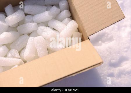 Close up of white expanded polystyrene foam pieces inside an open brown cardboard box placed on a surface of white snow. Stock Photo