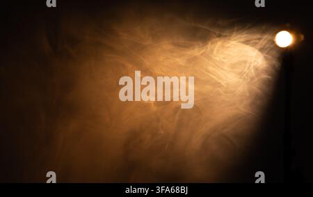 Dramatic warm spotlight beam shining through dense smoke on black background  Stock Photo