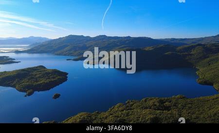 Stunning aerial view of a deep blue lake nestled among forested mountains, with small islands scattered across calm waters under a clear sky. Stock Photo