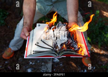 Hands hold an open book engulfed in bright orange flames with charred pages curling under the heat, symbolizing censorship, forbidden knowledge, and d Stock Photo