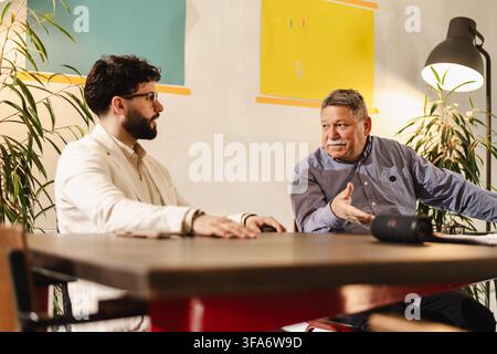 Two men engaged in discussion in a modern office setting with plants and colorful wall art during the day Stock Photo