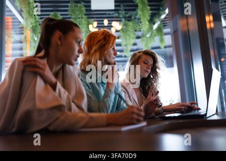 Businesswomen collaborating in a modern workspace while discussing ideas and reviewing documents on a laptop at a stylish cafe in the late afternoon Stock Photo