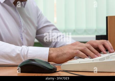 Close-up view shows a man in a white shirt typing on a computer keyboard at a desk. Stock Photo