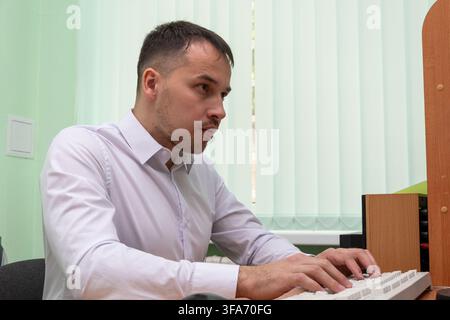 A focused young man in a white shirt types on a keyboard at an office desk. Stock Photo