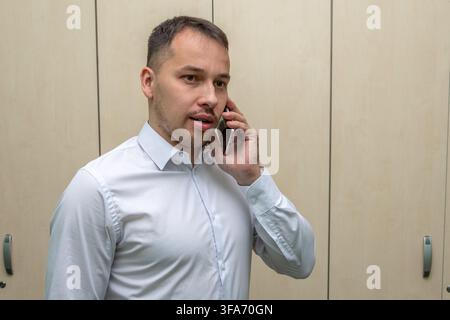 A young man in a white shirt speaks on a smartphone while standing indoors against light wooden cabinets. Stock Photo