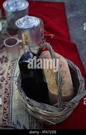A glass bottle of dark red wine and a loaf of homemade artisan sourdough bread in a woven basket. Rustic Greek countryside setup. Stock Photo