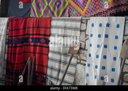 Display of vibrant, traditional woven Greek textiles, including red and white blankets and rugs with geometric patterns, hanging on a stone wall. Stock Photo