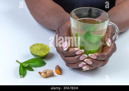 Two hands hold a glass mug containing a liquid and leaves. A lime half, mint leaves, and turmeric rhizomes are on the surface. Stock Photo