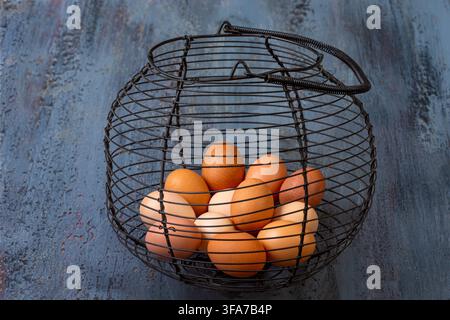 Brown and white chicken eggs are contained within a black wire basket resting on a dark, textured surface. Stock Photo
