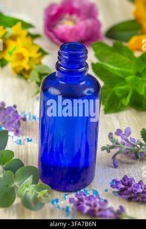 A blue glass bottle placed on a wooden surface, surrounded by various flowers and leaves, with some blue and purple petals scattered around. Stock Photo