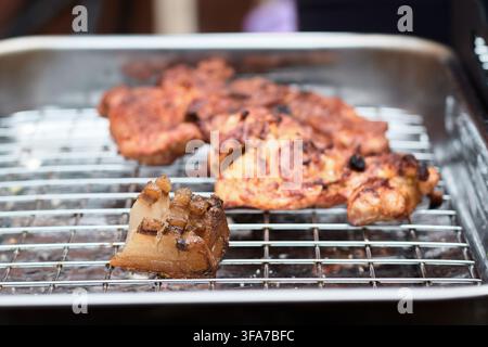Close-up of crispy pork lard pieces and seasoned roasted meat grilling on a metal wire rack inside a stainless steel roasting pan outdoors. Stock Photo