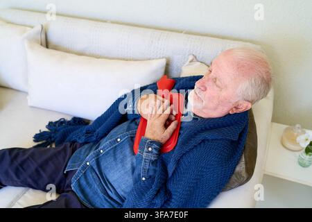 Senior man resting on a couch with a hot water bottle, home healthcare and elderly care for pain relief and comfort. Self care and independence in old Stock Photo