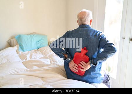 Senior man experiencing back pain, using a hot water bottle for self medication and comfort in a home healthcare setting. Elderly male patient seeking Stock Photo