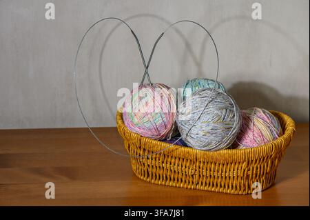A studio photograph featuring a wicker basket filled with several balls of multicolored speckled yarn, with circular knitting needles stuck into the y Stock Photo