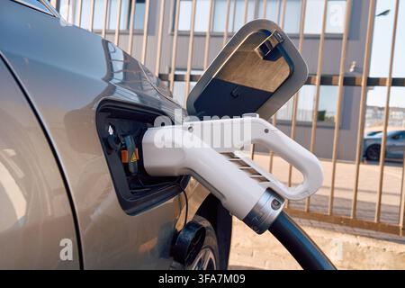 Electric vehicle charging plug connected to car at outdoor station with close view of port, cable, reflective body panel, and modern urban energy Stock Photo