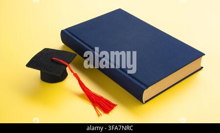 Graduation Cap Beside Dark Blue Book on Yellow Background Concept of Knowledge Academic Degree and Education Stock Photo