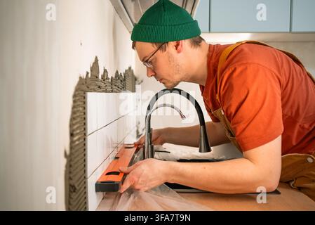 Professional tiler installing ceramic kitchen backsplash tiles, applying adhesive to a wall with a trowel, ensuring perfect alignment using a spirit level during home renovation work Stock Photo
