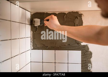 Worker spreading cement adhesive with a trowel while installing modern ceramic backsplash tiles in a kitchen during a home renovation, close-up on hands and wall progress Stock Photo