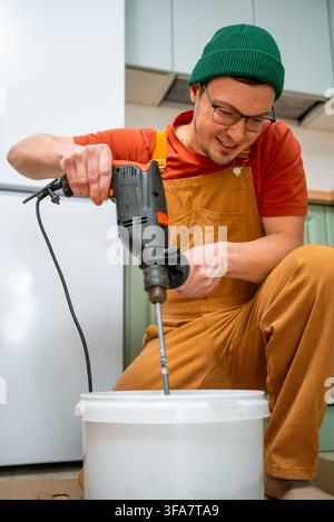 Man in orange overalls and green beanie smiling while mixing plaster or tile adhesive with an electric drill in a white bucket during a home renovation or diy repair project Stock Photo