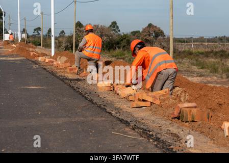 Construction workers laying bricks along the edge of a paved road at roadwork site Stock Photo