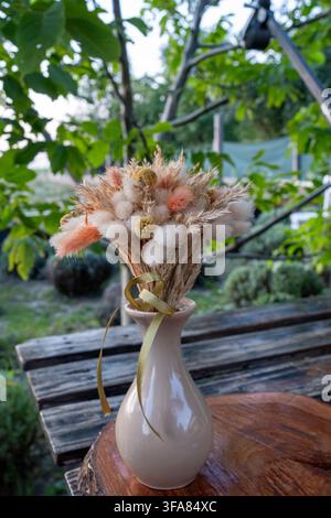 Fluffy botanical composition tied with ribbon sits inside glossy pottery vessel on timber surface outdoors under natural day light against blurred gre Stock Photo
