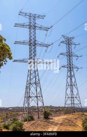 Two massive metal towers stand tall against a clear blue sky, supporting a network of high-voltage transmission lines over arid terrain Stock Photo