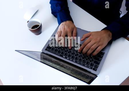 High angle flatlay view of a businessman in a dark blue shirt typing on a modern grey laptop keyboard, with a cup of coffee and a white computer mouse Stock Photo