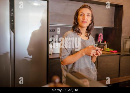 Woman standing in her kitchen and texting on a smartphone Stock Photo