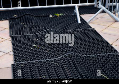 Perforated rubber safety mats with non-slip honeycombed surface texture laid on paving stones alongside steel crowd security barricades Stock Photo