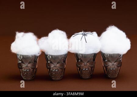 Four dark glass skull-shaped cups each topped with fluffy white cotton smoke, with a small black spider perched on one, arranged on a warm brown backg Stock Photo