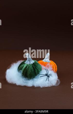 Ribbed green and orange velvet pumpkins resting on a fluffy white cotton cloud with a small black spider, set against a dark warm brown background. A Stock Photo