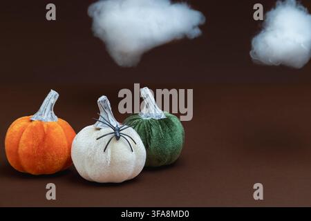 Orange, white, and green velvet pumpkins next to a small black spider, with two fluffy white cotton clouds floating above against a dark brown backgro Stock Photo