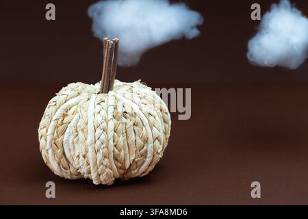 A handmade woven straw pumpkin with two wooden stem sticks, standing beneath two fluffy white cotton clouds floating against a dark brown background. Stock Photo