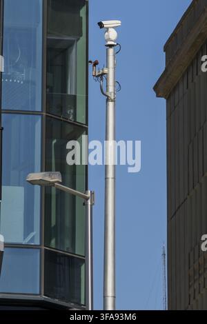 Security Surveillance Camera Mounted on Metal Pole Between Glass and Stone Buildings Stock Photo