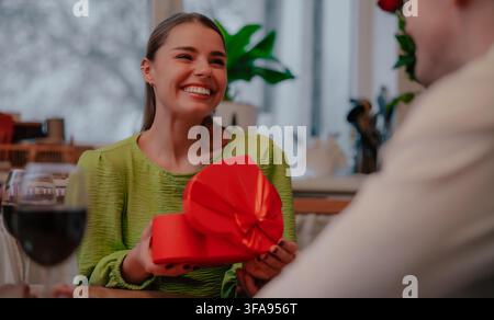Joyful woman laughs while holding an open red heart-shaped gift box at a romantic dinner table with her partner nearby. Stock Photo
