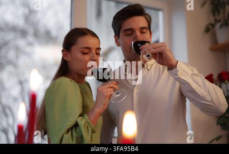 Young couple drinks red wine together beside glowing candles during an intimate romantic evening in a cozy home kitchen. Stock Photo