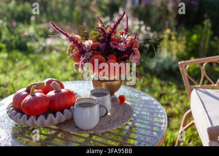 Hot coffee served in backyard on table by bouquet of fall flowers, pumpkins in morning autumnal garden. Lounge furniture. Drinks for two in peaceful s Stock Photo