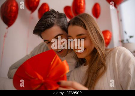 Excited young couple leans together and smiles down into a red heart-shaped gift box surrounded by red heart balloons. Stock Photo