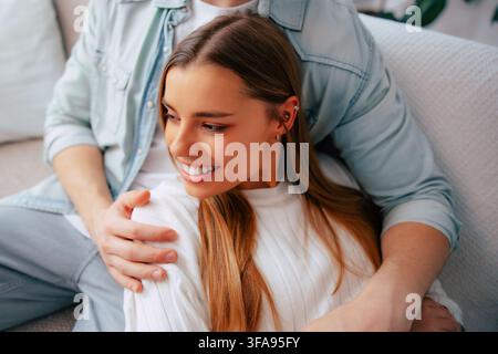Young woman smiles while sitting comfortably in her partner's arms on a sofa in a bright modern home interior. Stock Photo
